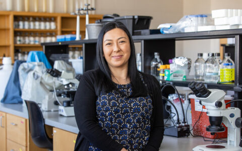 Biological science researcher stands in a lab with microscopes. DoD award recipient.