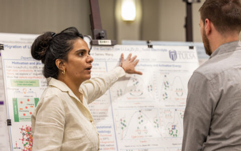 Physics doctoral student presenting battery research at UTulsa, explaining charts on a poster board.