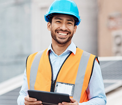 Photograph of a smailing man wearing an orange safety vest and blue hardhat while holding a clipboard