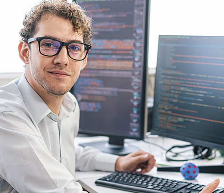 Photograph of a man with curly hair and glasses sitting at a desk with two computer monitors displaying lines of text
